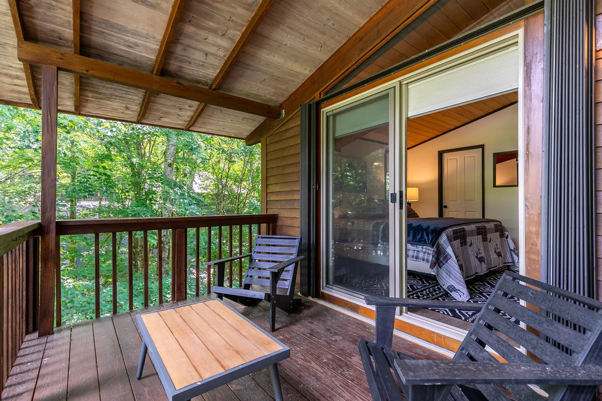 An exterior scene focusing on a comfortable outdoor deck with casual furniture, looking out to a serene view of mature trees. The sliding glass door hints at the easy movement between indoor comfort and outdoor exploration. The mood is peaceful and inviting, ideal for unwinding with a pet after a walk.