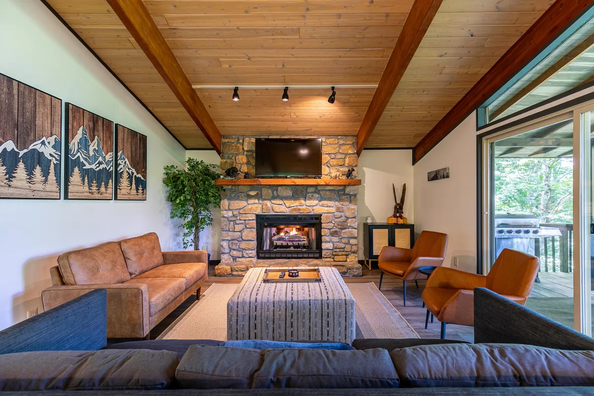 A cozy interior living area with a crackling fireplace, plush sofas, a coffee table, and large windows letting in natural light. The atmosphere is warm, inviting, and ideal for relaxation or apres-ski moments.