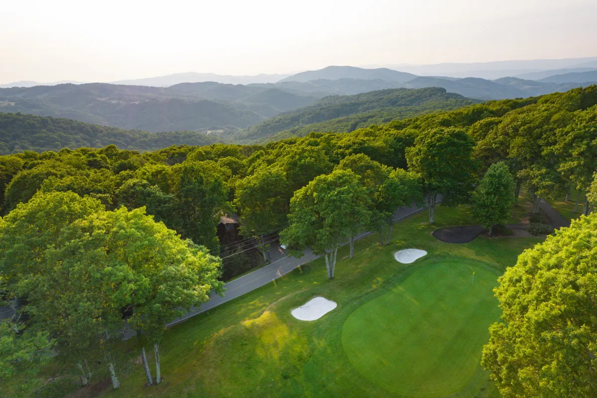 A panoramic view of the Beech Mountain Club's golf course set against a backdrop of a mountain range and lush greenery. The perfectly maintained fairways stretch into the distance, with natural beauty on all sides, and the soft morning light creates a tranquil atmosphere.