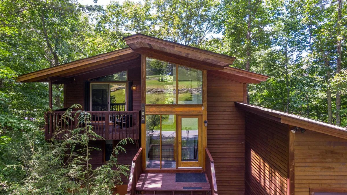 A welcoming exterior view of the home featuring large glass windows, a wooden front deck, and an asymmetrical roofline. People are seen gathering on the deck, surrounded by lush greenery, hinting at the home's connection to nature and group outdoor experiences.