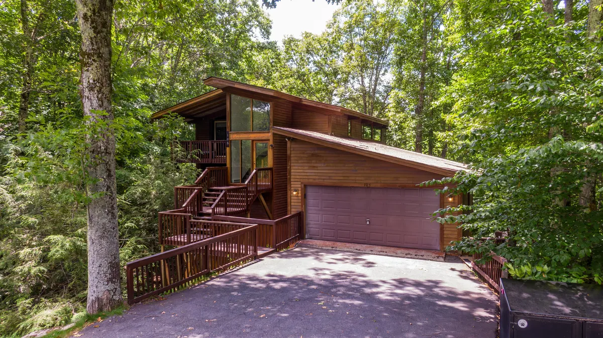 The image showcases the inviting wooden exterior of a recently renovated single-family house nestled among tall trees. The spacious deck, accented by large windows, is perfectly positioned to capture sunset views and invites guests to relax outdoors, surrounded by nature. Stairs lead up to the home, emphasizing easy access and a welcoming entryway.