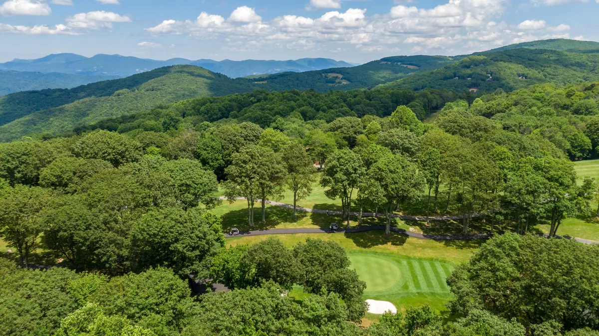 An inviting, wide-angle perspective of the golf course framed by rolling hills, lush green trees, and distant mountains. The scene is peaceful, with ample open space and hints of wildlife habitat, suggesting both recreational and natural opportunities.
