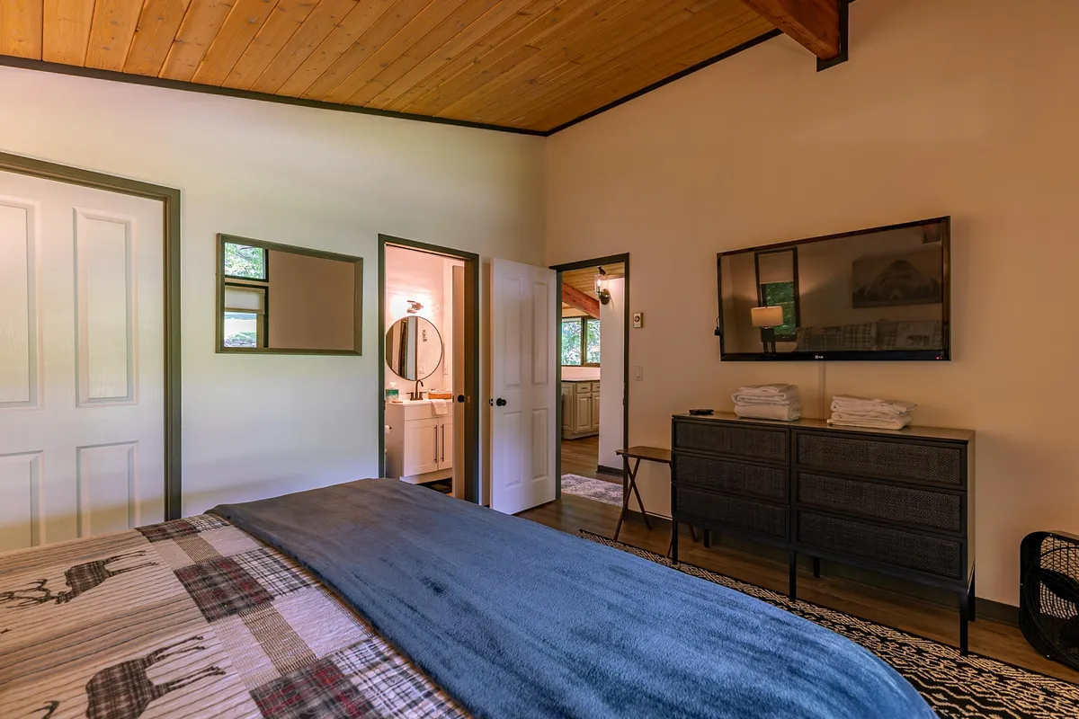 A comfortable bedroom with plaid bedding, a wooden ceiling, a wall-mounted TV, a dressing table with a mirror, and an open door revealing a glimpse of the bathroom beyond. Natural light brightens the room, highlighting its cozy and welcoming appeal.