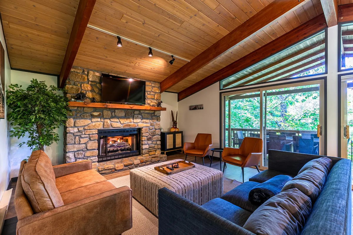 This interior view showcases the great room with a striking stone fireplace as the centerpiece, surrounded by plush seating. The space is filled with natural light streaming through expansive floor-to-ceiling windows, while wooden beams overhead add rustic mountain charm. The arrangement of furnishings invites social interaction and relaxation.