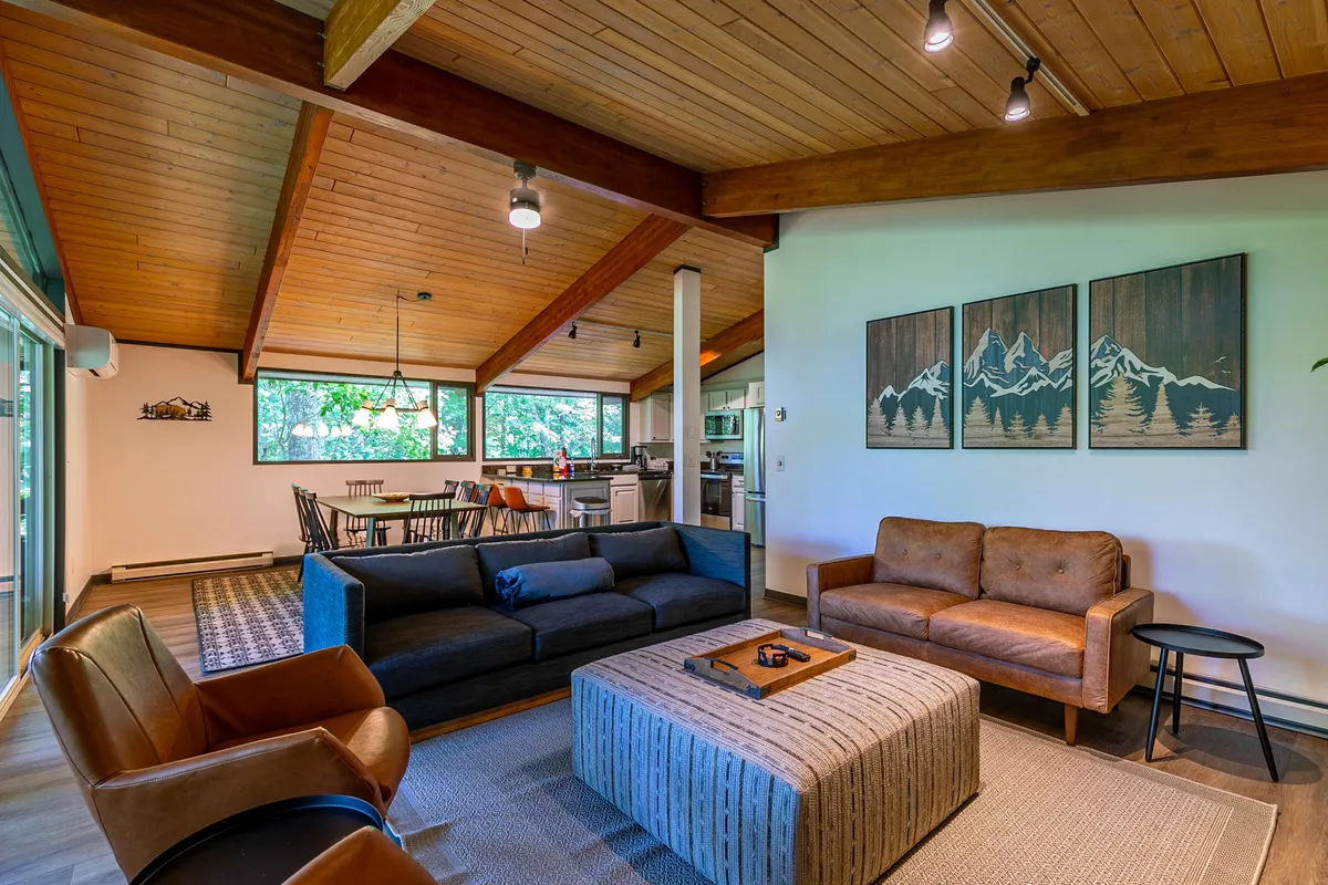 An open-plan living-dining area featuring comfortable sofas, a central coffee table, wall art, and a dining table visible in the background. The space is brightened by large windows that offer serene woodland views, emphasizing easy flow and communal atmosphere.