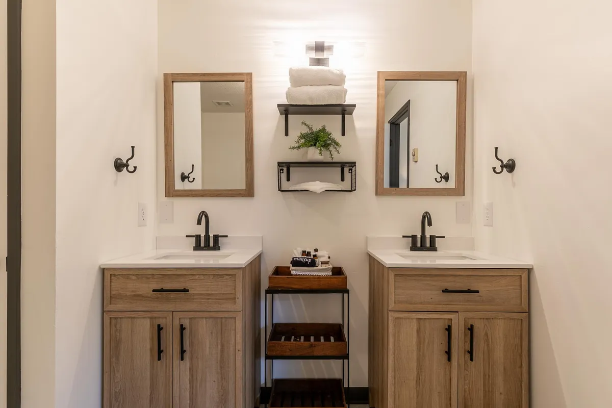A modern bathroom featuring double vanity sinks, large mirrors, wooden shelving with neatly arranged towels, and organized storage for toiletries. The ambiance is clean, inviting, and spa-like.