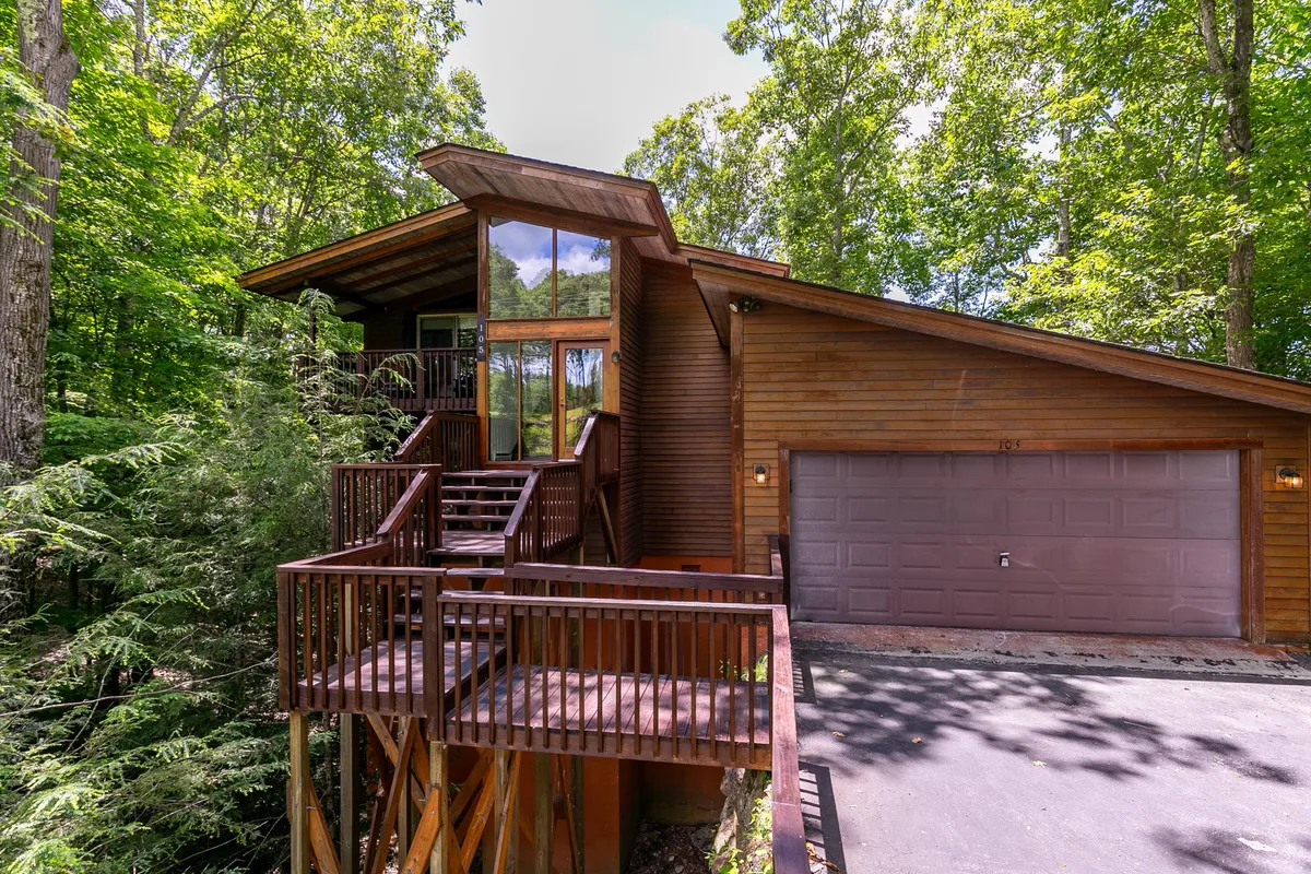 A tranquil exterior view of the chalet showcasing a spacious wooden deck surrounded by lush greenery, with large windows and a distinctive sloped roof visible. The deck appears inviting for relaxation and overlooks natural trails leading into the forest, emphasizing easy access to outdoor adventures right from the home.